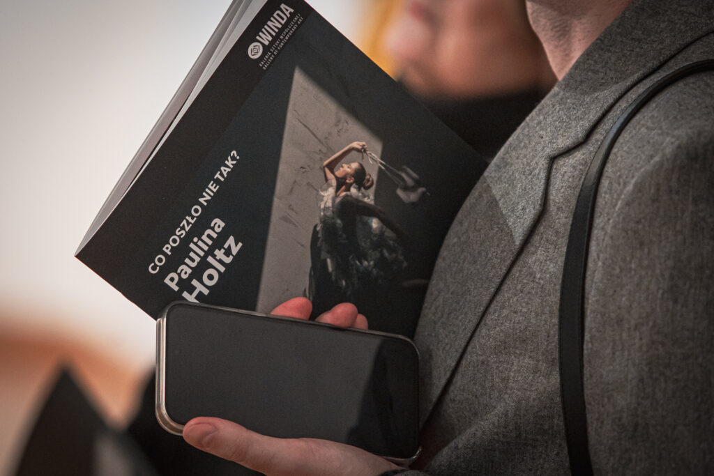 Zdjęcie. Katalog wystawy Paulina Holtz "Coś poszło nie tak". Katalog trzyma człowiek ubrany w szarą marynarkę.