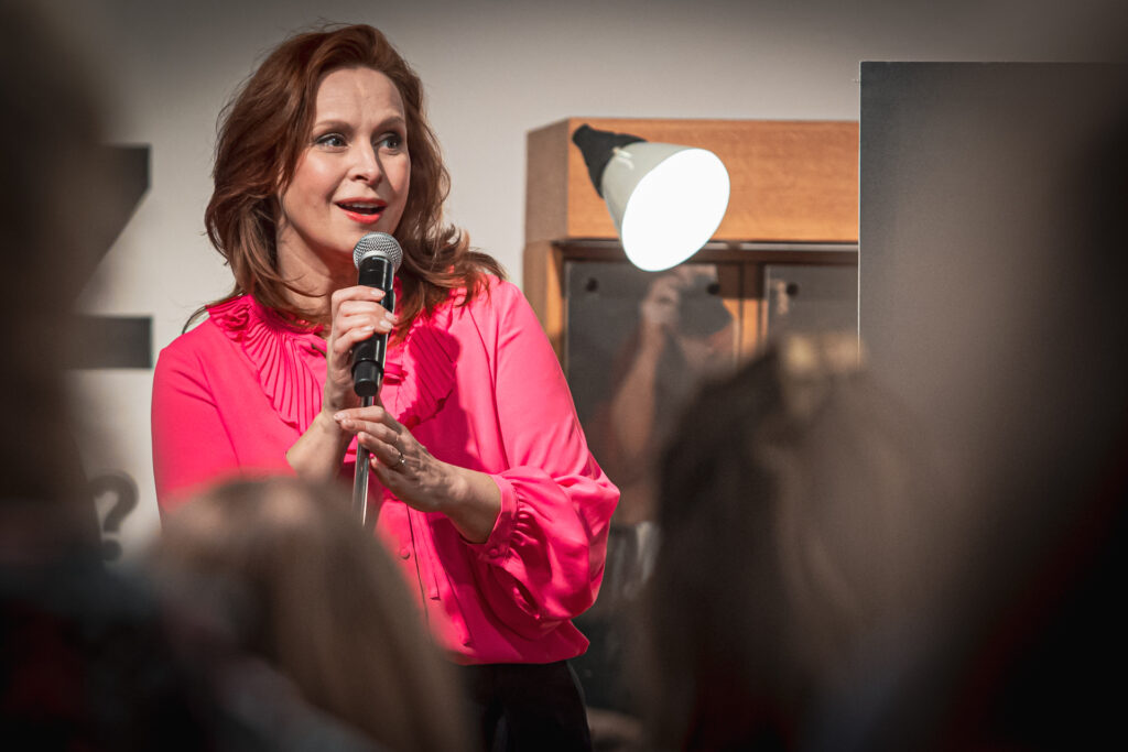Zdjęcie. Rudowłosa kobieta mówi do mikrofonu. To aktorka Paulina Holtz podczas wernisażu wystawy swoich fotografii.