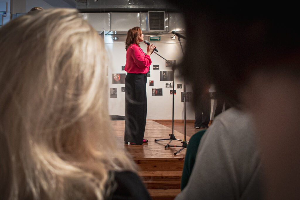 Zdjęcie. Kobieta w czarnych spodniach i różowej bluzce mówi do mikrofonu. Przed sceną stoją widzowie wernisażu fotografii Pauliny Holtz.