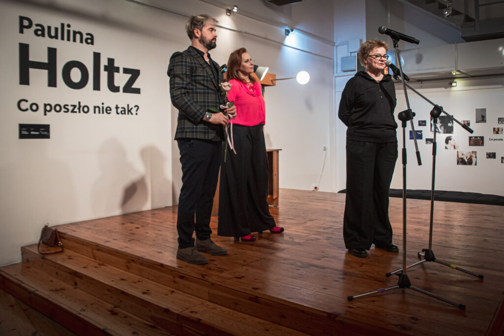 Zdjęcie. Trójka ludzi na podeście podczas wernisażu. Do mikrofonu mówi kobieta ubrana na czarno. Z tyłu stoją Paulina Holtz i Artur Wijata.