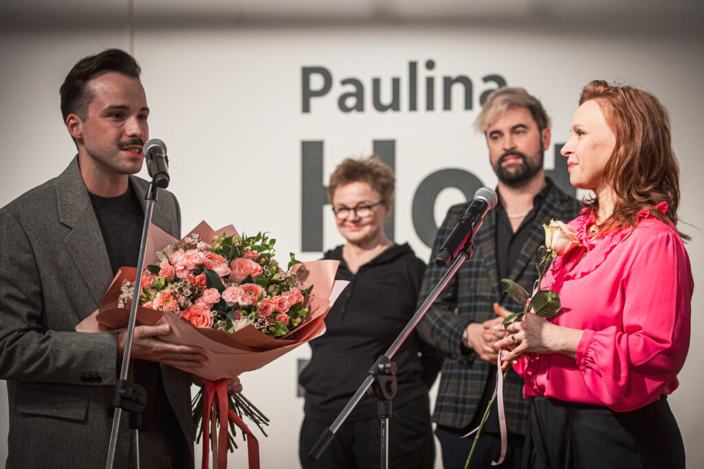 Zdjęcie. Szczupły mężczyzna z wąsami i bukietem rózowych kwiatów mówi do mikrofonu, patrząc na Paulinę Holtz.