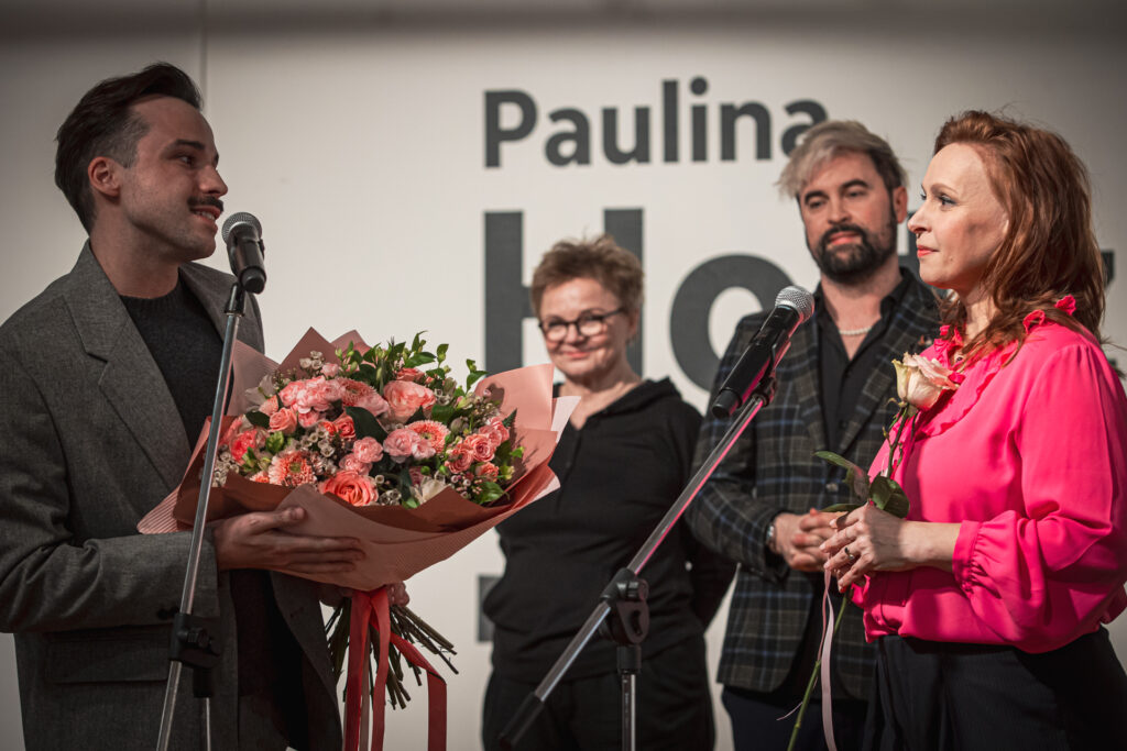 Zdjęcie. Szczupły mężczyzna z wąsami i bukietem rózowych kwiatów mówi do mikrofonu, patrząc na Paulinę Holtz.