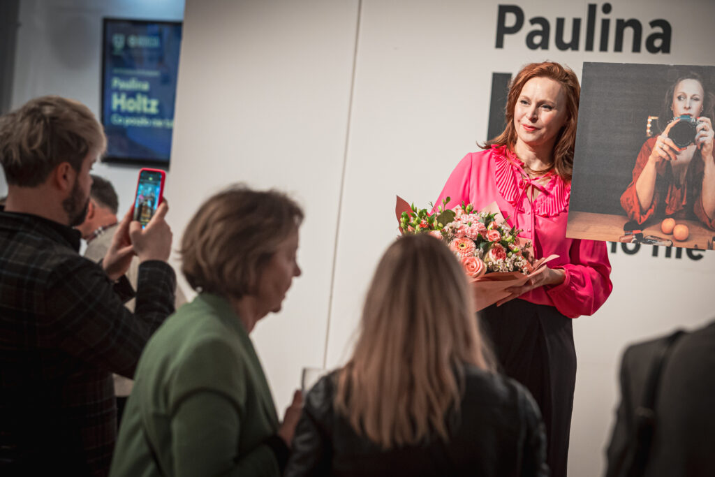 Zdjęcie. Kobieta w intensywnie różowej bluzce z kołnierzem i bukietem kwiatów w dłoniach stoi na podwyższeniu. To Paulina Holtz, autorka zdjęć w Galerii Winda. Przed nią inni ludzie, goście wernisażu.