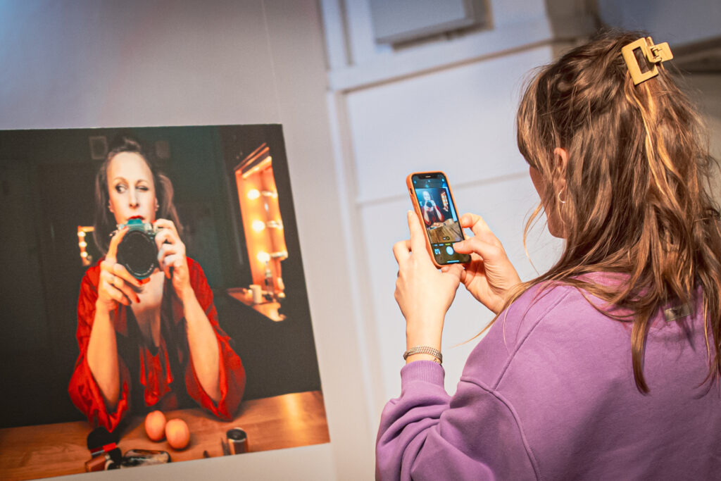 Zdjęcie. Kobieta w fioletowej bluzie ustawia telefon do zdjęcia. Fotografuje planszę z reprodukcją zdjęcia przedstawiającego Paulinę Holtz w czerwonej bluzce, trzymającej w dłoniach aparat fotograficzny.