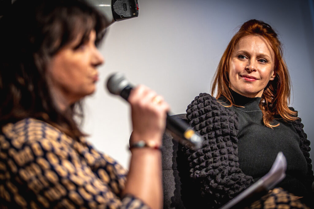 Zdjęcie. Uśmiechnięta ruda kobieta przygląda się brunetce mówiącej do mikrofonu. To Paulina Holtz i prowadząca spotkanie Magdalena Fudala.