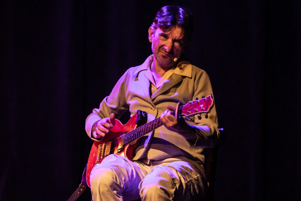 Zdjęcie. Mężczyzna gra na gitarze ze skrzywioną miną.