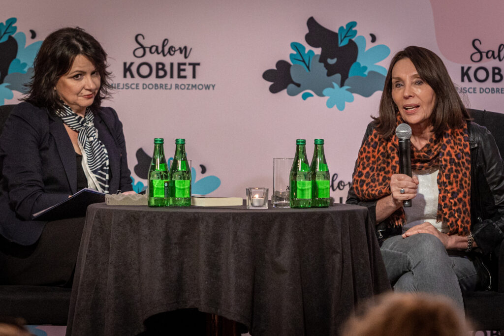 Zdjęcie. Salon Kobiet z Ewą Ewart. Dwie kobiety siedzą przy stoliku. Brunetka w ciemnej marynarce i czarno-białej apaszce na szyi patrzy w kierunku widowni. Druga szczupła brunetka o prostych włosach do ramion z apaszką w motyw centek w kolorze rudo-czarnym mówi do mikrofonu. To prowadząca spotkanie Magdalena Fudala i gościni Salonu Kobiet Ewa Ewart.