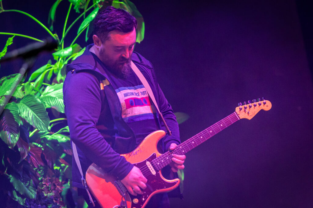 Zdjęcie. Mężczyzna z brodą w bluzie z kapturem gra na gitarze.