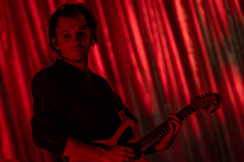 Zdjęcie. Mężczyzna o dłuższych włosach gra na gitarze. Za nim czerwona opuszczona kurtyna.