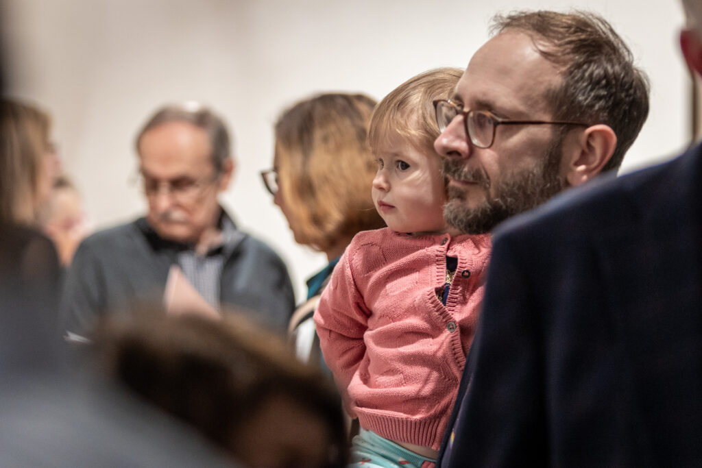 Zdjęcie. Szczupły mężczyzna z brodą i w okularach trzyma na rękach kilkuletnią dziewczynę w różowym sweterku. Obok inni ludzie.