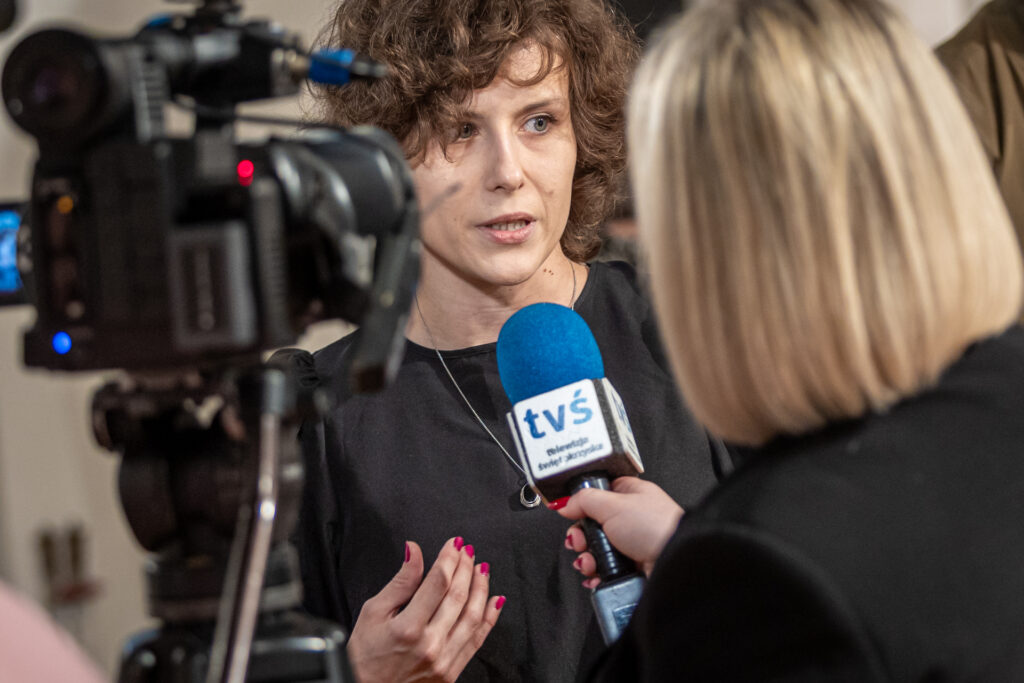 Zdjęcie. Młoda kobieta o włosach kręconych do ramion mówi do mikrofonu z logo Telewizji Świętokrzyskiej. Mikrofon trzyma blondynka o prostych wlosach do ramion. Obok kamera telewizyjna.