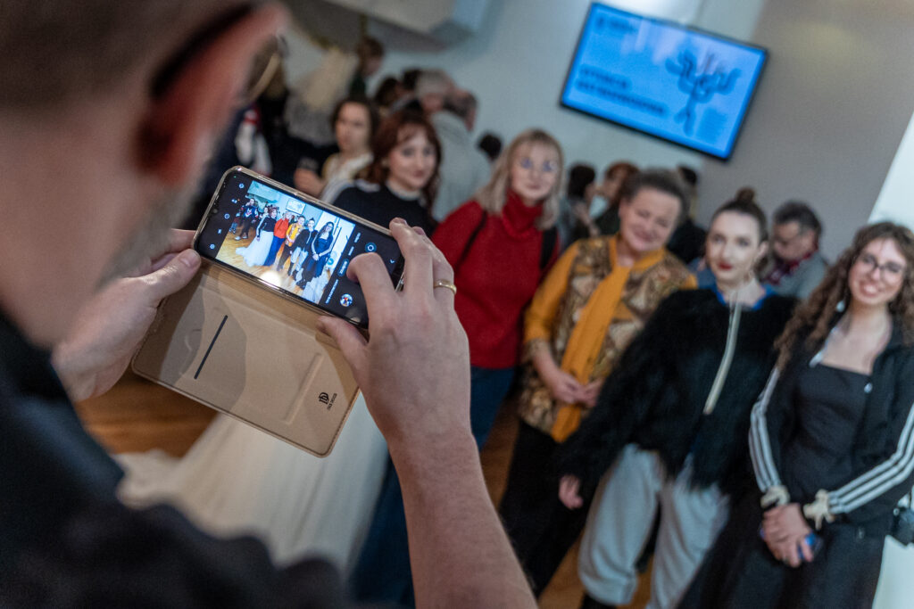 Zdjęcie. Wernisaż wystawy "Sytuacja jest rozwojowa". Grupa ludzi pozuje do zdjęcia, które mężczyzna robi swoim smartfonem.
