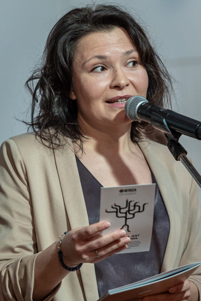 Zdjęcie. Wernisaż wystawy "Sytuacja jest rozwojowa". Brunetka w beżowej marynarce mówi do mikrofonu. To Daria Maroń-Ptak.