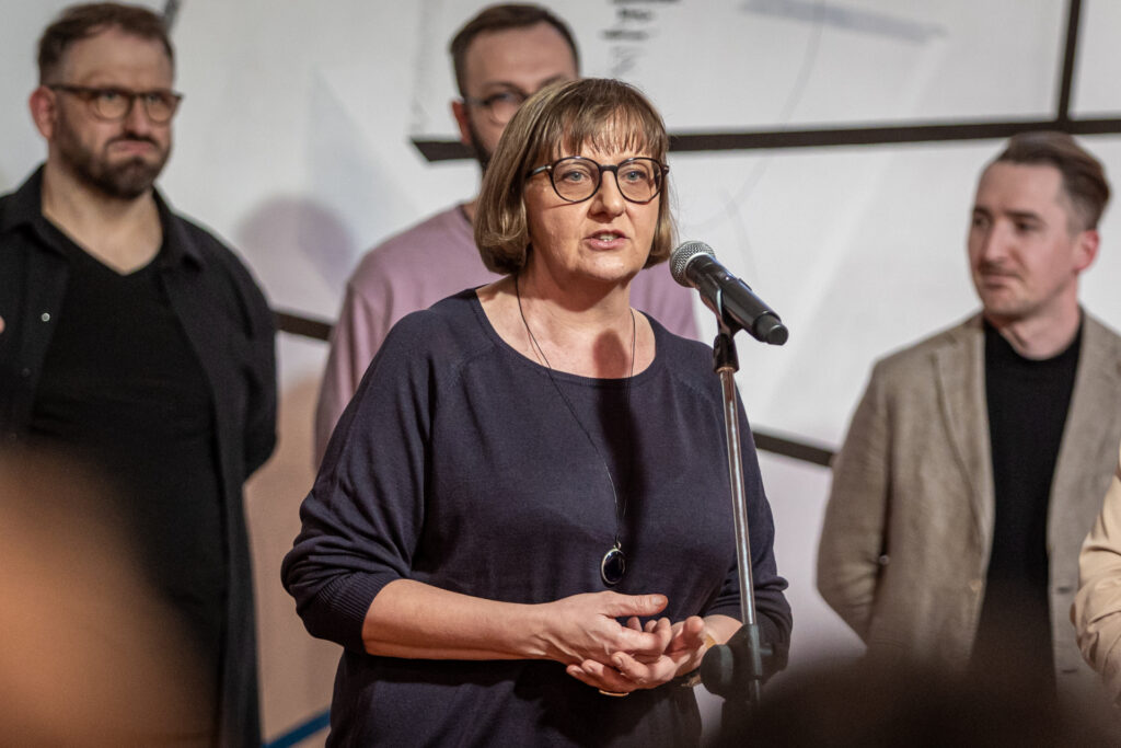 Zdjęcie. Wernisaż wystawy "Sytuacja jest rozwojowa". Przy mikrofonie stoi kobieta o jasnych prostych włosach do ramion i dużych okularach w ciemnych oprawkach. To Iwona Stachura.