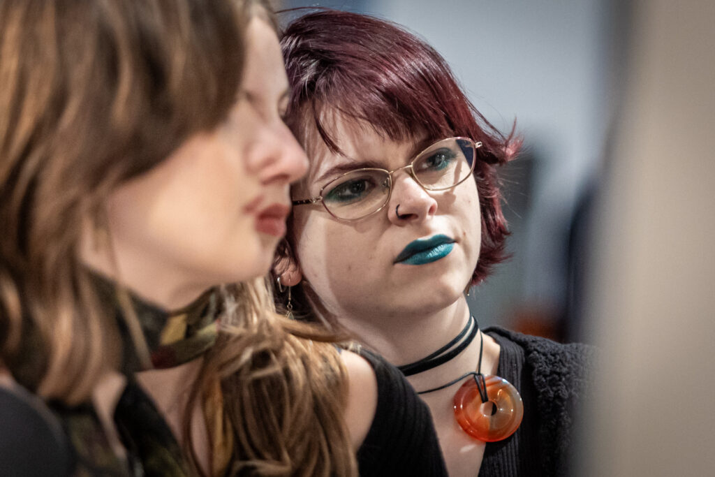 Zdjęcie. Wernisaż wystawy Dyplom. Młode dziewczyny patrzą w kierunku pracy. Jedna z nich ma rubinowe włosy i na zielono umalowane usta.