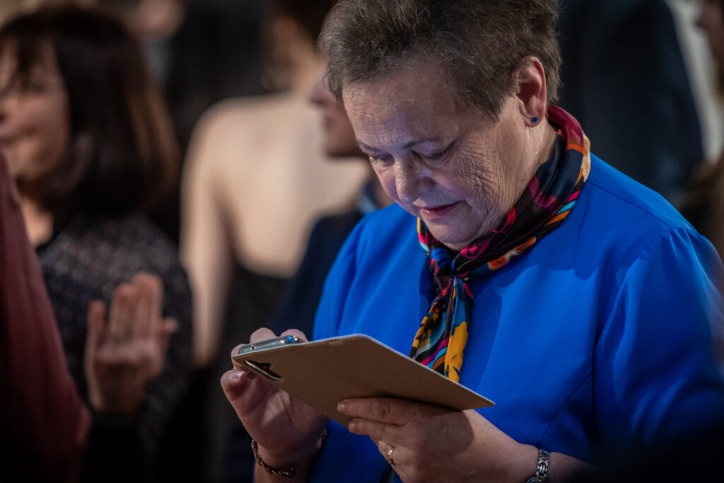 Zdjęcie. Wernisaż wystawy Dyplom. Starsza kobieta w niebieskiej sukience z kolorową apaszką na szyi patrzy w ekran telefonu komórkowego.