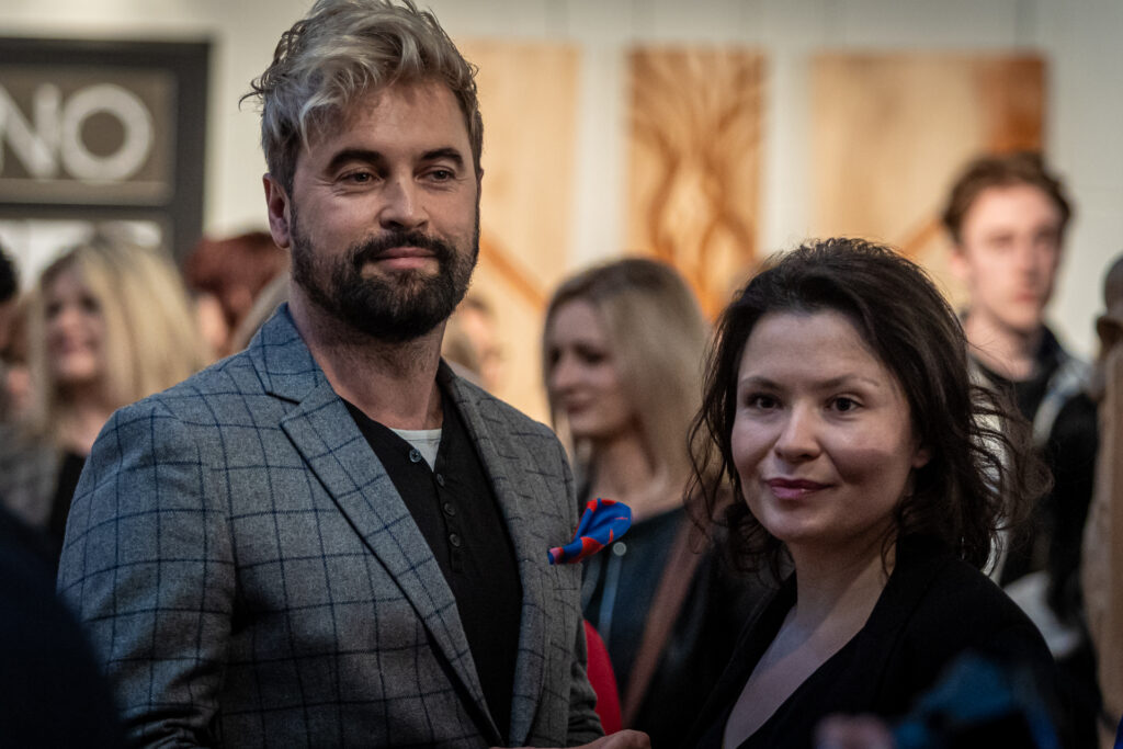 Zdjęcie. Wernisaż wystawy Dyplom. Wysoki mężczyzna z jasną grzywką i ciemną brodą patrzy przedd siebie uśmiechając się. To Artur Wijata. Obok niewysoka brunetka. To Daria Maroń-Ptak.