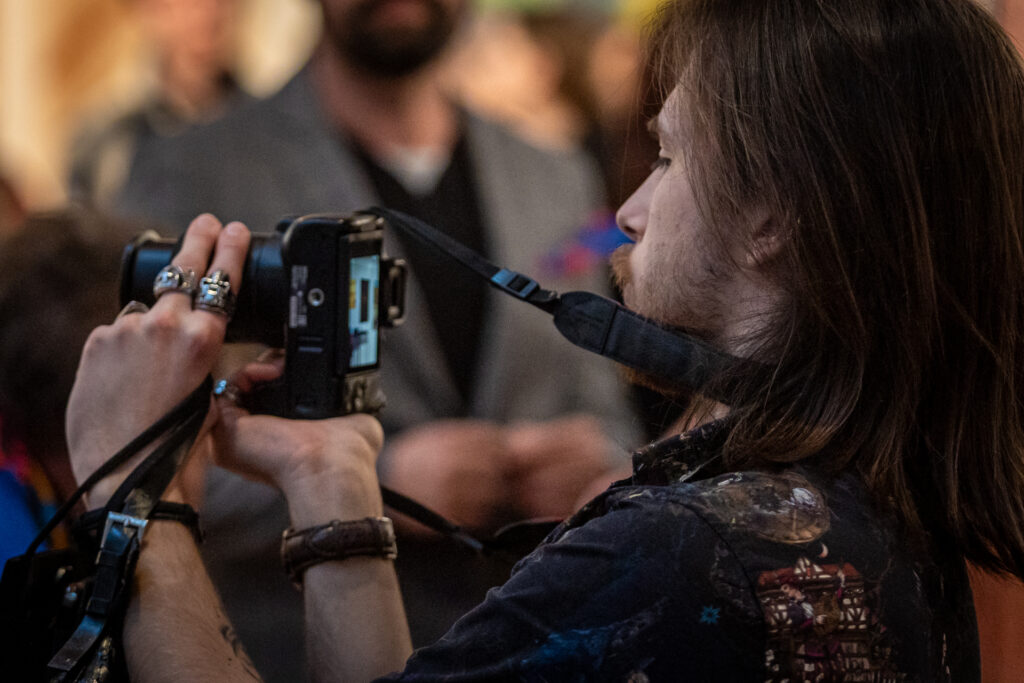 Zdjęcie. Mężczyzna z dłuższymi ciemnymi włosami i brodą patrzy na wyświetlacz aparatu fotograficznego. Szykuje się do zrobienia zdjęcia.