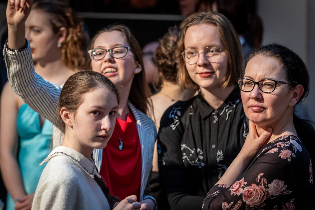 Grafika. Grupa młodych dziewczyn stoi blisko siebie. Obok nich pani w średnim wieku w ciemnych oprawkach i kwiecistej sukience. Patrzą przed siebie w różną stronę.