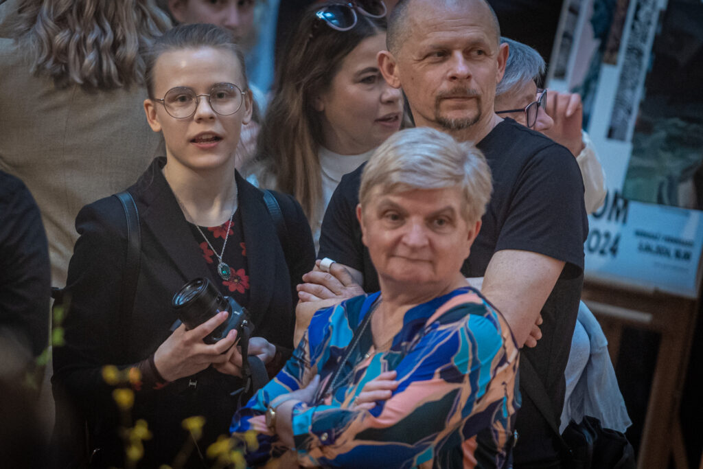 Zdjęcie. Wernisaż wystawy Dyplom. Trzy osoby: starsza kobieta blondynka, łysawy mężczyzna brodą i młoda dziewczyna w okularach z aparatem fotograficznym w ręku patrzą przed siebie.