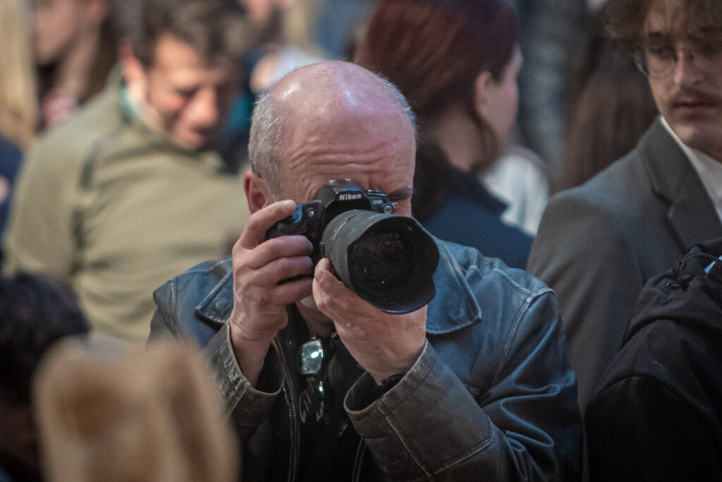Zdjęcie. Łysawy mężczyzna robi zdjęcie aparatem marki Nikon.