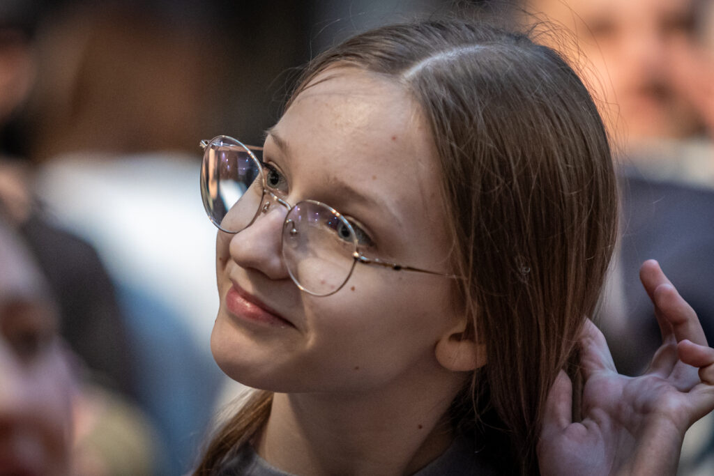 Zdjęcie. Wernisaż wystawy Dyplom. Młoda dziewczyna o długich włosach z przedziałkiem na środku głowy i okularach w metalowych oprawkach patrzy przed siebie, delikatnie się uśmiechając.