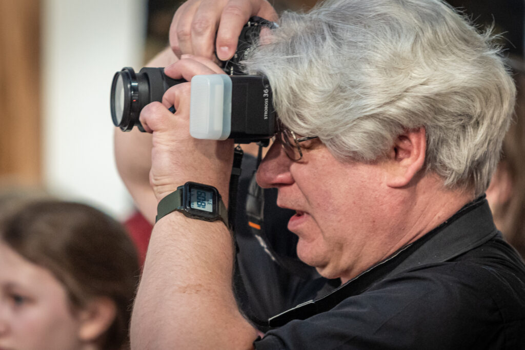 Zdjęcie. Siwy mężczyzna o dłuższych włosach przystawia aparat fotograficzny do oka, robiąc zdjęcie.