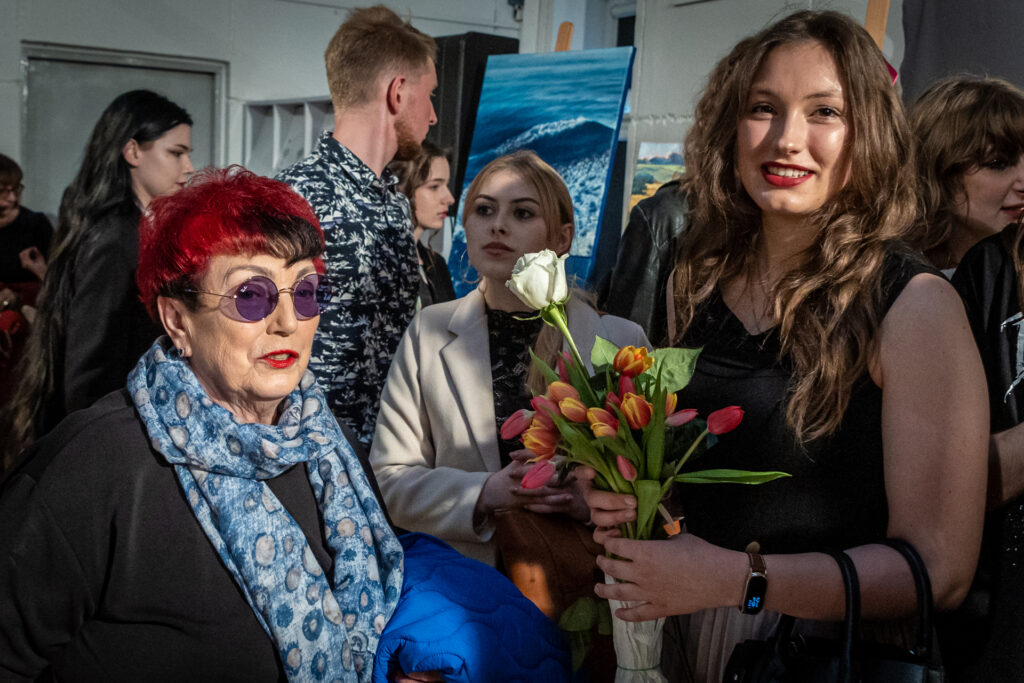 Zdjęcie. Młoda dziewczyna w czarnej sukience pozuje do zdjęcia ze starszą panią o o czerwonawych włosach. Dziewczyna trzyma w dłoniach bukiet tulipanów.