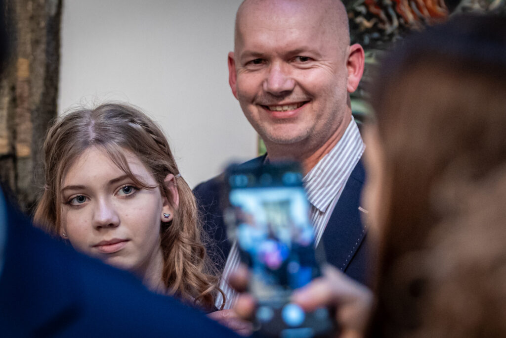 Zdjęci. Starszy łysy mężczyzna i młoda dziewczyna o blond włosach pozują do zdjęcia, które ktoś robi telefonem komórkowym.