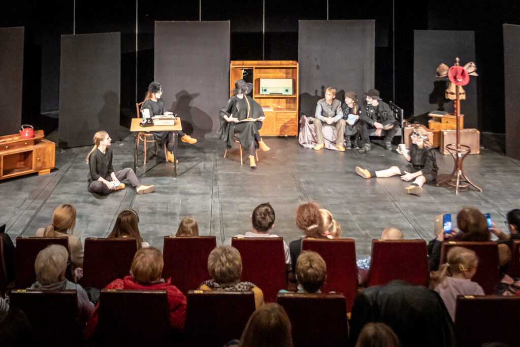 Zdjęcie. Mała Scena KCK podczas spektaklu Studia Pantomimy. Na scenie siedmiu aktorów. W centralnym miejscu na krześle siedzi dziewczyna w dużym kapeluszu. Dookoła niej pozostali aktorzy. Wszyscy patrzą w jej kierunku.