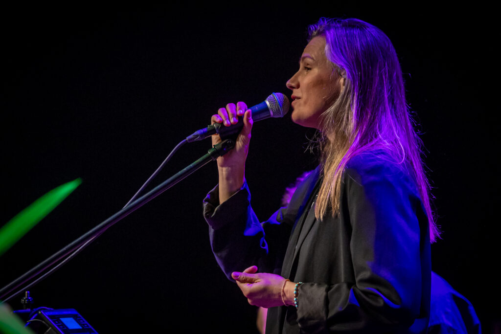 Zdjęcie. Blondynka o długich prostych włosach w ciemnej marynarce śpiewa do mikrofonu. To Natalia Grosiak, wokalistka zespołu Mikromusic.