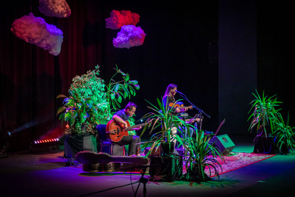 Zdjęcie. Duża Scena KCK. Na scenie trio Mikromusic. W centralnym miejscu wokalistka śpiewa do mikroonu. Po obu stronach na krzesłach siedzą gitarzyści. W tle zielona ściana roślin. Nad muzykami chmurki oświetlone kolorowymi reflektorami.