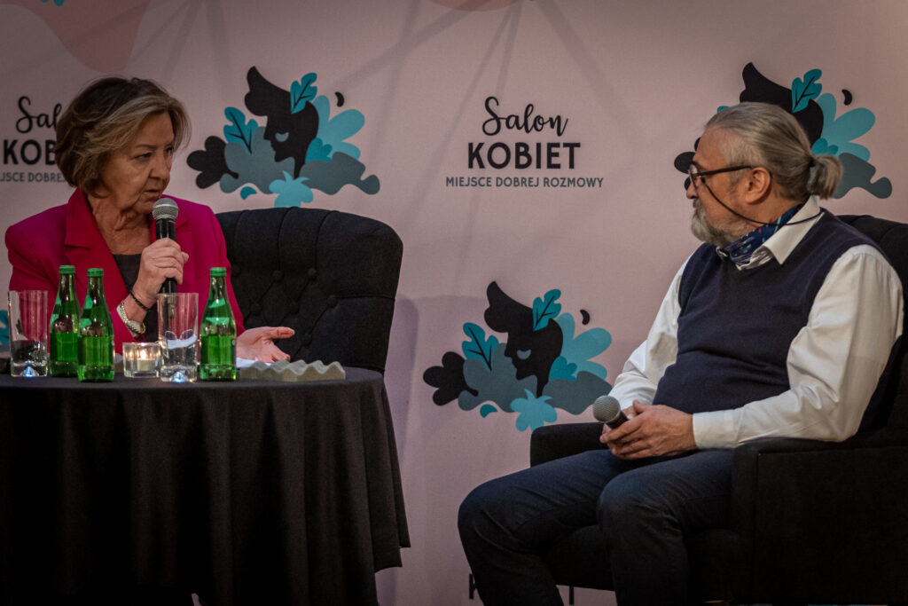 Zdjęcie. Dwójka ludzi siedzi przy okrągłym stoliku. W tle duża ścianka z napisem Salon Kobiet. Kobieta patrzy na mężczyznę i jednocześnie mówi do mikrofonu. Mężczyzna patrzy na kobietę. To Marzena Sobala prowadząca spotkanie i gość Andrzej Kruszewicz, dyrektor warszawskiego ZOO.