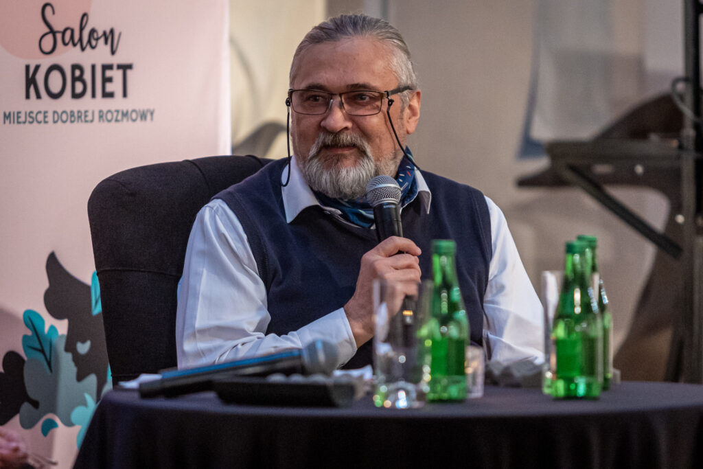 Zdjęcie. Siwy mężczyzna z brodą i w okularach z uśmiechem mówi do mikrofonu. To dyrektor warszawskiego ZOO Andrzej Kruszewicz. Patrzy w bok.