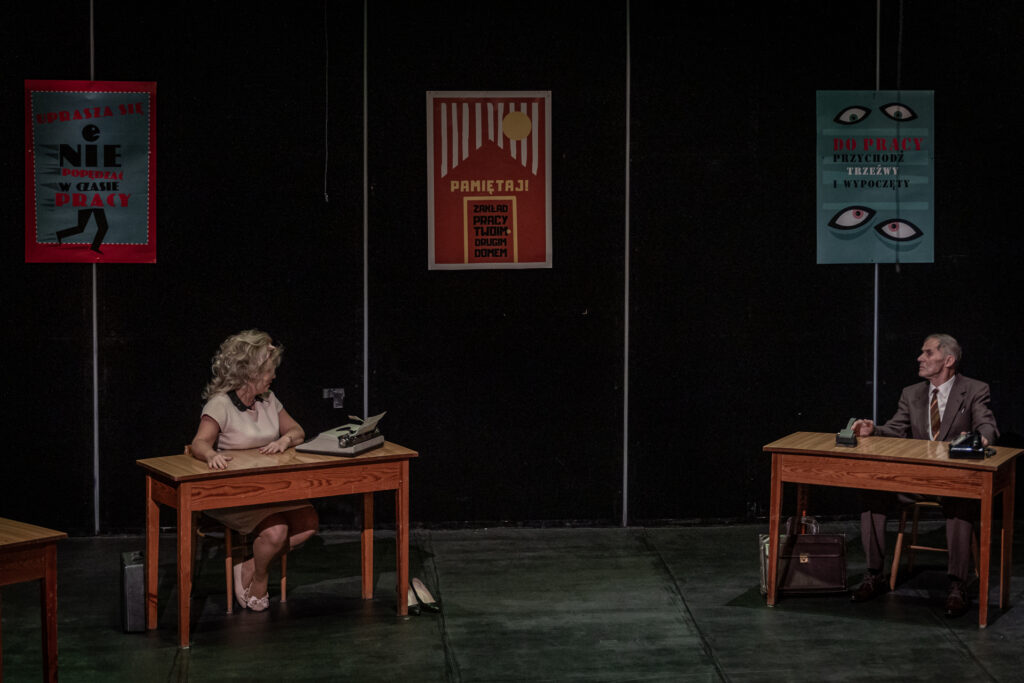 Zdjęcie. Występ Teatru bez Suflera. Na scenie dwa biurka, przy których siedzą kobieta i mężczyzna. Kobieta jest blondynką o utapirowanej, pełnej loków fryzurze. Mężczyzna ma na sobie garnitur. Patrzą na siebie.