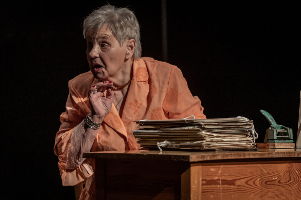 Zdjęcie. Spektakl Teatru bez Suflera. Starsza kobieta o krótkich ufryzowanych włosach siedzi za biurkiem. Odwraca się w bok i mówi coś.