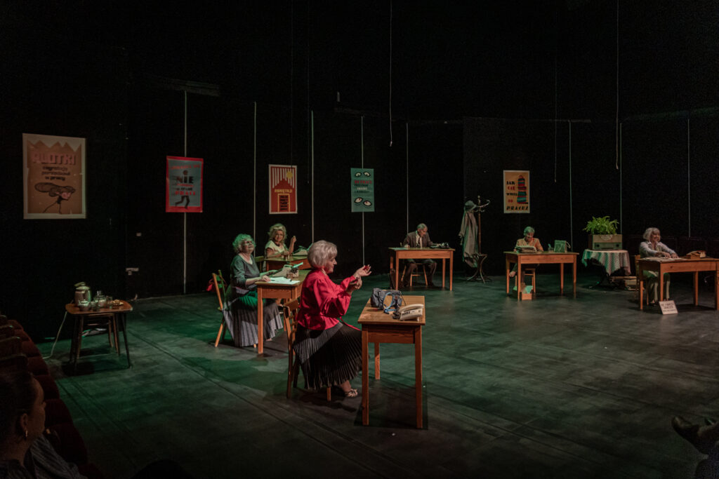 Zdjęcie. Spektakl teatru bez Suflera. Mała Scena KCK. Na scenie sześć biurek, przy każdym siedzi jedna osoba: pięć kobiet i jeden mężczyzna. W tle duże plakaty.