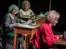 Zdjęcie. Trzy kobiety siedzą przy biurkach. Dwie z tyłu patrzą w kierunku pierwszej. Środkowa uśmiecha się. Na burku ma papiery, szklankę w metalowym koszyczku i maszynę do pisania.