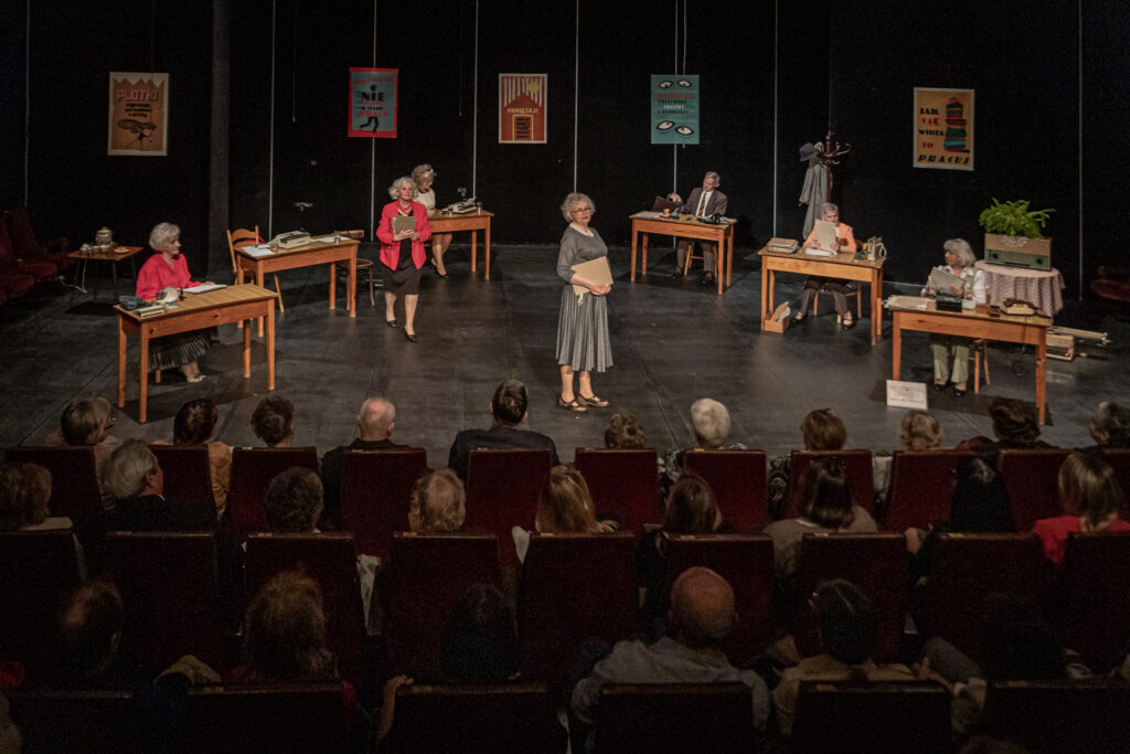 Zdjęcie. Scena ze spektaklu Czekoladki dla prezesa Teatru bez Suflera. Na środku stoi kobieta w szarej sukience z teczką od pachą. Przy biurkach pozostali aktorzy.