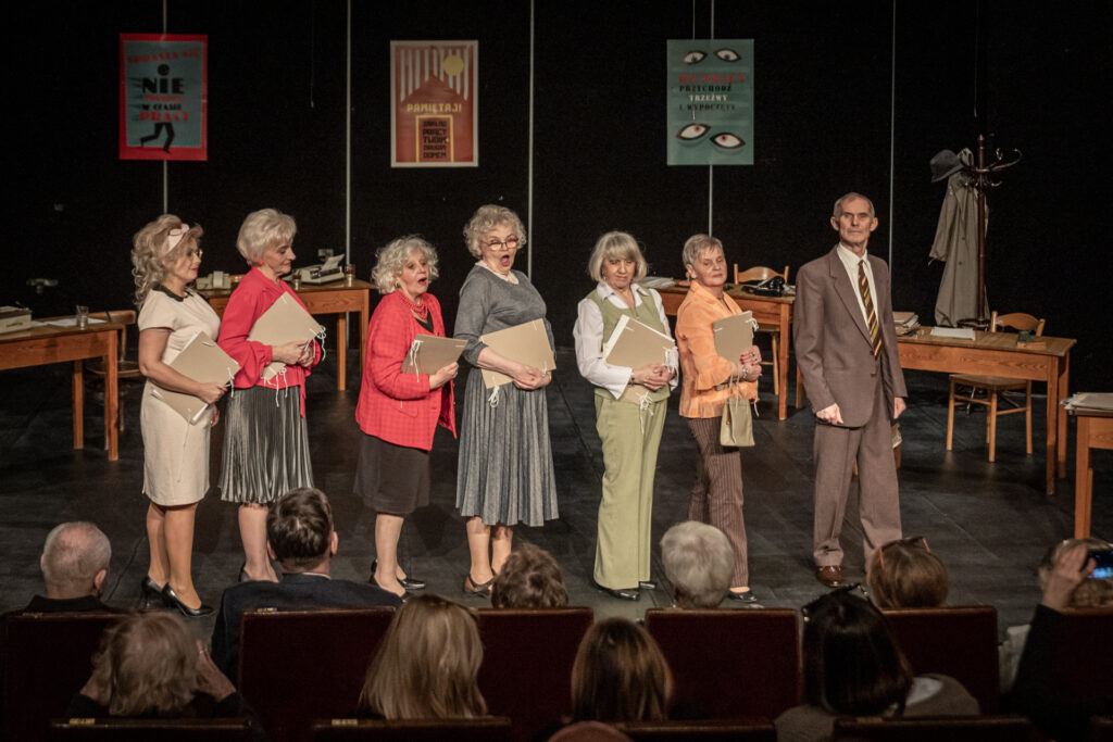 Zdjęcie. Zespól Teatru bez suflera stoi w rzędzie prawym bokiem do widowni, na przodu sceny. Wszyscy pod pachą ściskają teczki z dokumentami.