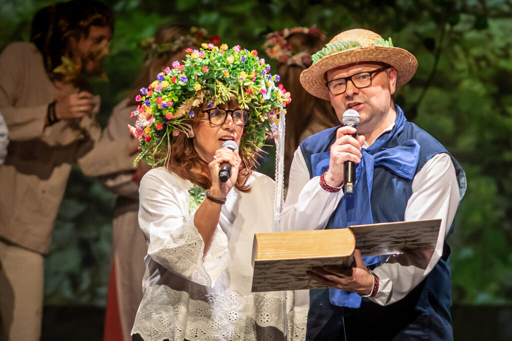 Zdjęcie. Para ludzi: kobieta w ogromnym wianku na głowie i mężczyzna w słomkowym kapeluszu mówią do mikrofonów. Mężczyzna trzyma przed obojgiem ogromną otwartą księgę.