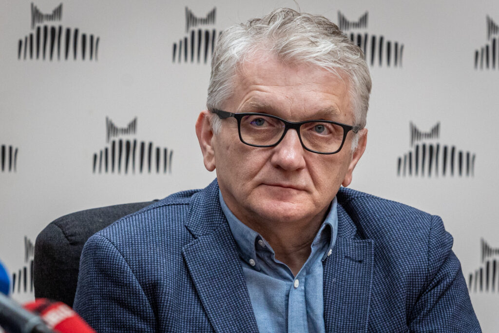 Zdjęcie. Mężczyzna o siwych włosach w okularach o cienkich czarnych oprawkach w niebieskiej koszuli marynarce w granatowo-czarną drobną kratę patrzy przed siebie. To aktor Dariusz Kowalski, który wcielał się w rolę Józefa Tracza w serialu "Plebania".