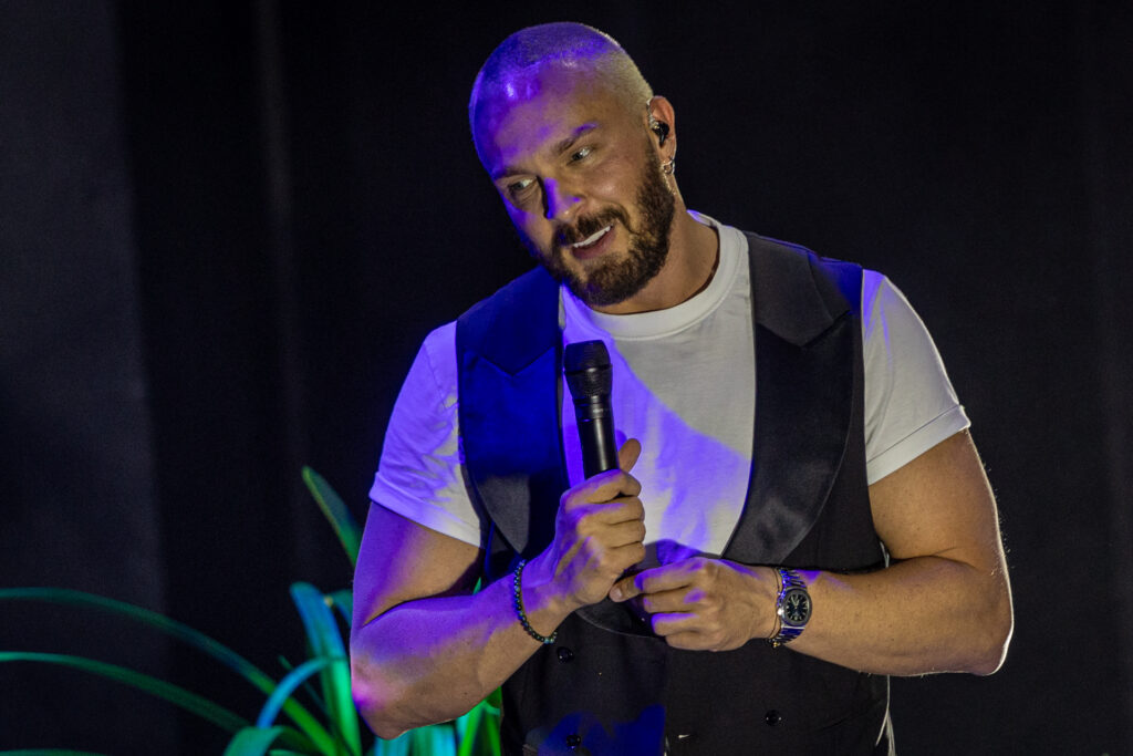 Zdjęcie. Uśmiechnięty mężczyzna w białej podkoszulce i czarnej kamizelce patrzy przed siebie opuszczając głowę w bok. Uśmiecha się.