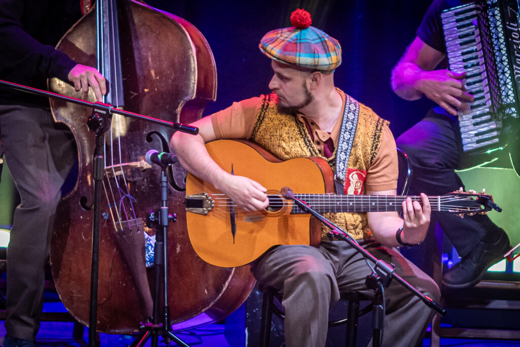 Zdjęcie. Warszawskie Combo Taneczne na scenie. Gitarzysta gra na gitarze. Ma na sobie kamizelkę z syrenką warszawską a na głowie kraciasty beret z daszkiem i czerwonym pomponem na czubku.