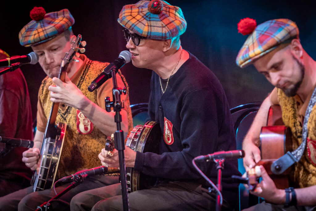 Zdjęcie. Trzech mężczyzn w kraciastych beretach z czerwonymi pomponami siedzi na krzesłach. Jeden z nich gra na bandżo, drugi na gitarze. Mężczyzna w środku gra na bandżo i śpiewa. To Jan Młynarski.