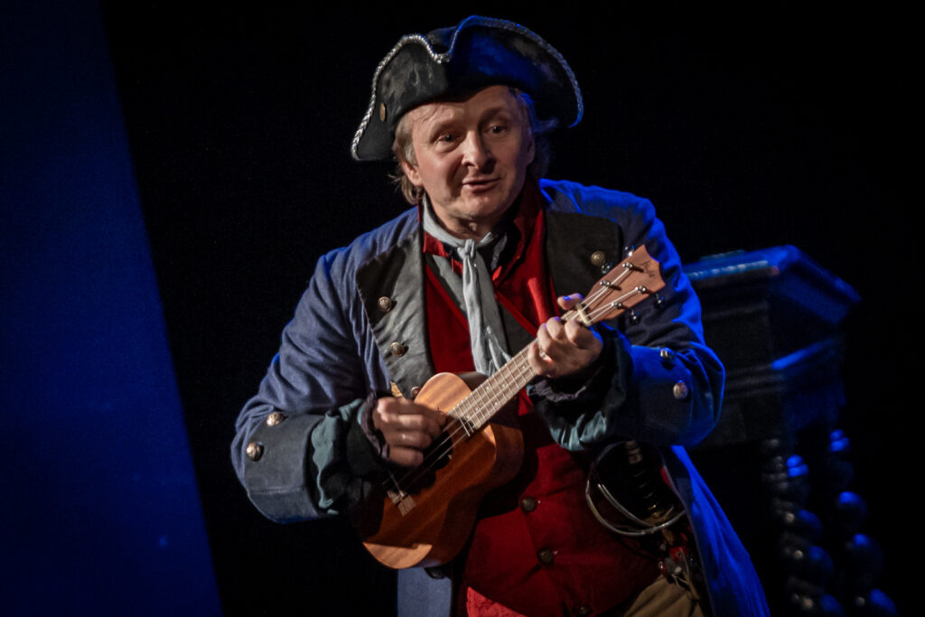 Zdjęcie. Mężczyzna w trójrożnej czapce na głowie, w czerwonej kamizelce i ciemnym kaftanie gra na małej gitarze. To Papkin z "Zemsty".