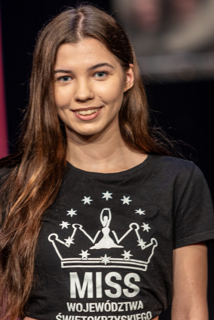 Zdjęcie. Młoda dziewczyna o długich włosach uśmiecha się patrząc przed siebie. Jest szatynką w czarnej podkoszulce z napisem: miss województwa świętokrzyskiego.
