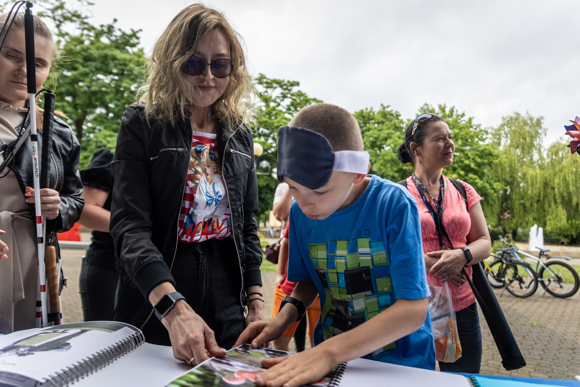 Zdjęcie. Chłopiec z czarną opaską podniesioną na czoło dotyka książki z fotografiami zwierząt. Nad nim stoi dziewczyna z długimi pofalowanymi włosami w ciemnych okularach na oczach.