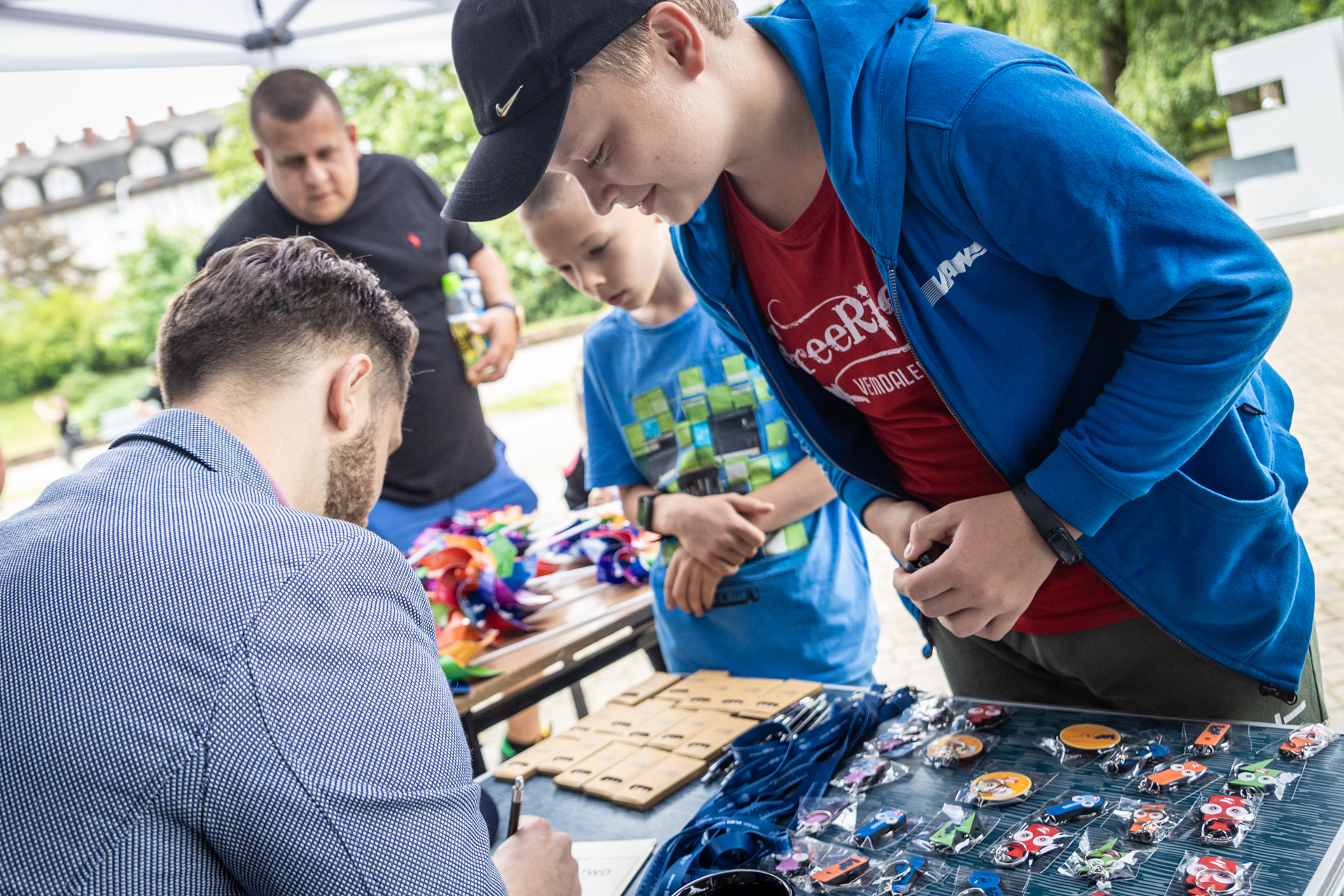 Zdjęcie. Chłopak w czarnej bejsbolówce i niebieskiej bluzie z kapturem pochyla się nad mężczyzną w garniturze, który coś pisze.