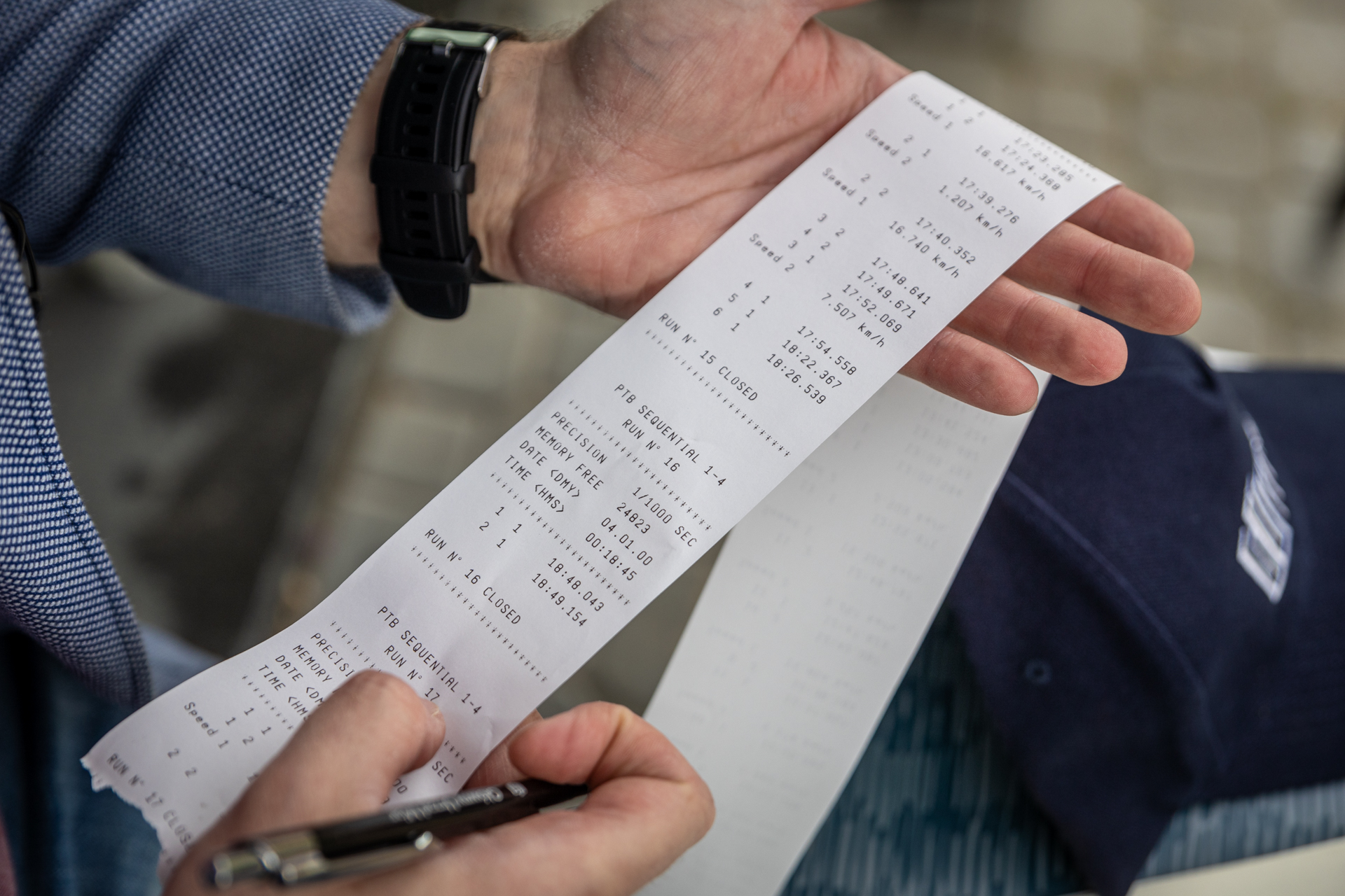 Zdjęcie. Męskie dłonie prezentujące długi pasek papieru wyglądający jak paragon z cyframi i napisami.